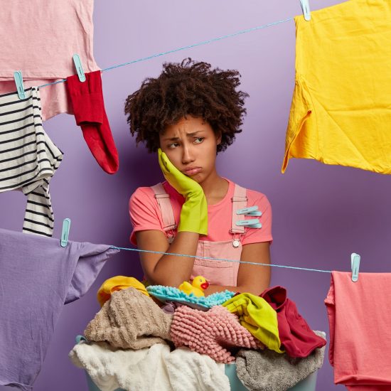 Upset tired Afro woman busy with housework, wears rubber gloves, dries clothes, has much jobs around house, stands near basket of dirty linen, isolated over purple background. Laundry day concept